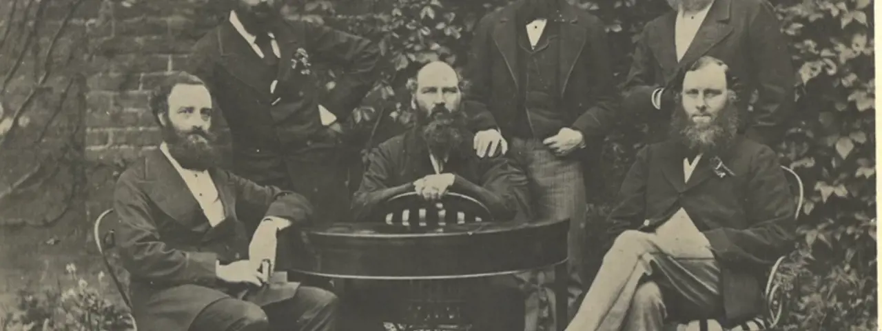 Black and white photo of six bearded men gathered around a table dressed in formal suits from the 1800s, threee seated and three standing